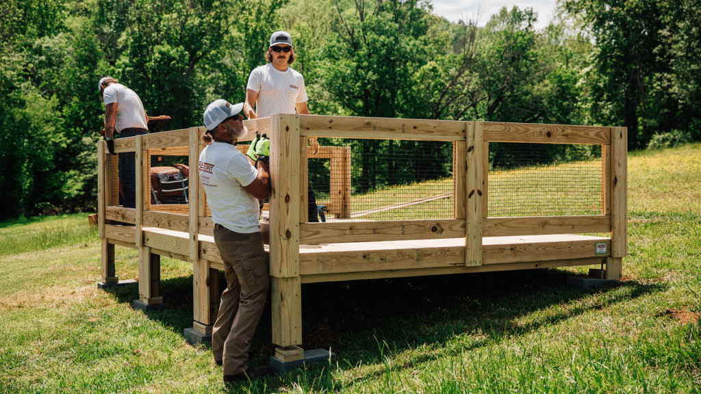 A group of workers wearing gloves and safety gear are assembling a raised wooden platform with railings in a grassy outdoor area surrounded by trees on a sunny day.