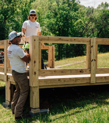 A group of workers wearing gloves and safety gear are assembling a raised wooden platform with railings in a grassy outdoor area surrounded by trees on a sunny day.