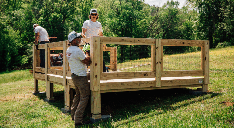 A group of workers wearing gloves and safety gear are assembling a raised wooden platform with railings in a grassy outdoor area surrounded by trees on a sunny day.