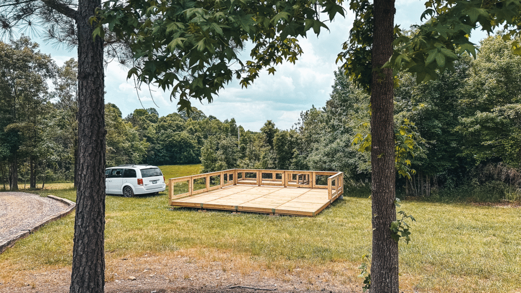 A wooden platform under construction on a grassy field surrounded by trees, with a white minivan parked nearby on a gravel path. The scene suggests a campground development in a natural, forested area.