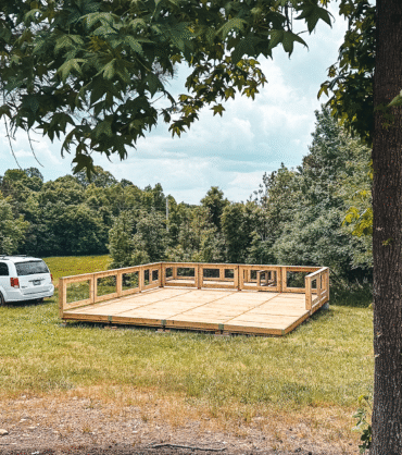 A wooden platform under construction on a grassy field surrounded by trees, with a white minivan parked nearby on a gravel path. The scene suggests a campground development in a natural, forested area.