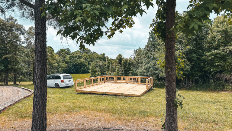 A wooden platform under construction on a grassy field surrounded by trees, with a white minivan parked nearby on a gravel path. The scene suggests a campground development in a natural, forested area.