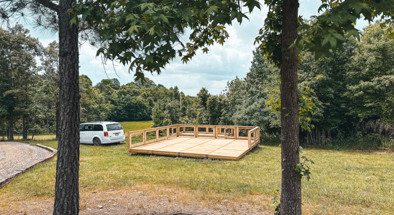A wooden platform under construction on a grassy field surrounded by trees, with a white minivan parked nearby on a gravel path. The scene suggests a campground development in a natural, forested area.