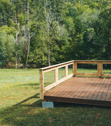 A wooden platform with a partial railing stands on a grassy field surrounded by dense, green forest. The platform appears to be elevated slightly off the ground and is supported by concrete blocks. The scene is bright and sunny, with a backdrop of tall trees and a clear, peaceful natural environment.