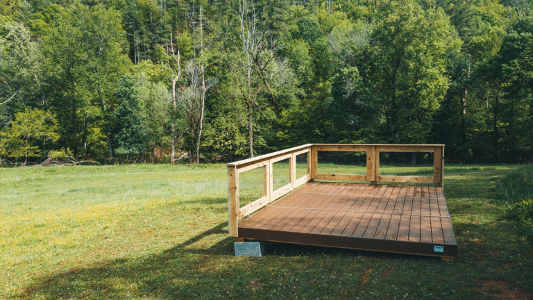 A wooden platform with a partial railing stands on a grassy field surrounded by dense, green forest. The platform appears to be elevated slightly off the ground and is supported by concrete blocks. The scene is bright and sunny, with a backdrop of tall trees and a clear, peaceful natural environment.