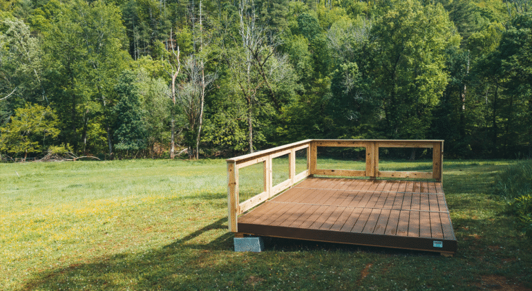 A wooden platform with a partial railing stands on a grassy field surrounded by dense, green forest. The platform appears to be elevated slightly off the ground and is supported by concrete blocks. The scene is bright and sunny, with a backdrop of tall trees and a clear, peaceful natural environment.