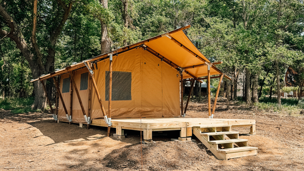 A safari-style glamping tent with a raised wooden platform and stairs, set in a forested area with tall trees and natural surroundings.