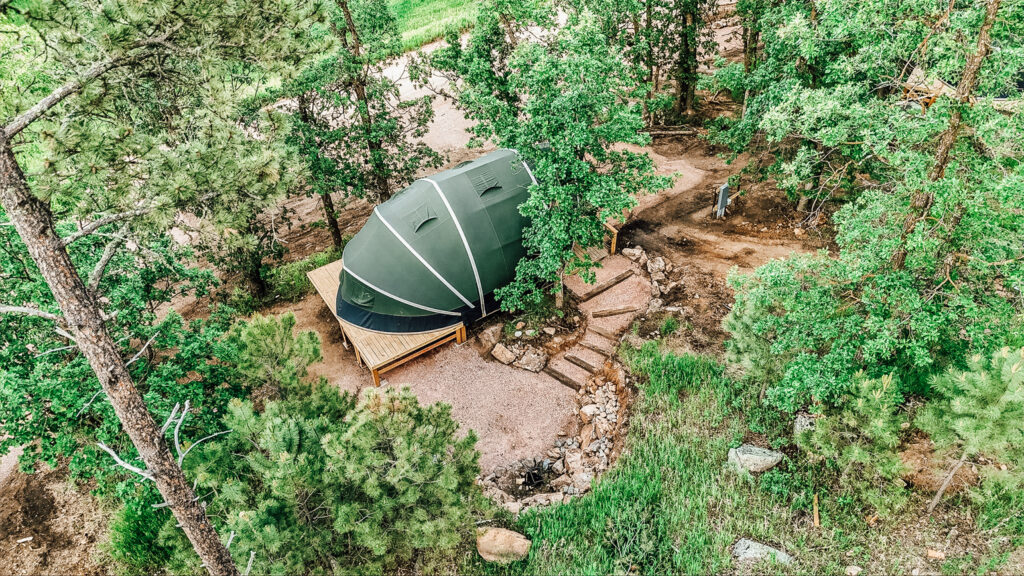 Aerial view of a modern glamping dome tent set on a raised wooden deck in a forested area, surrounded by trees, stone pathways, and natural greenery.