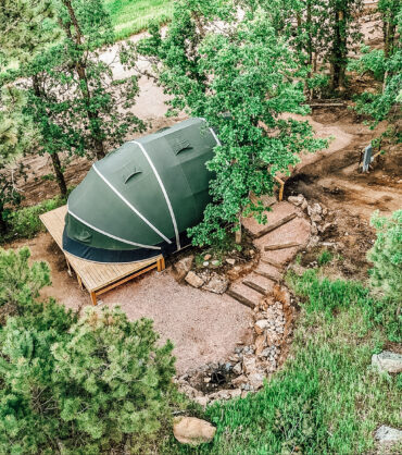 Aerial view of a modern glamping dome tent set on a raised wooden deck in a forested area, surrounded by trees, stone pathways, and natural greenery.