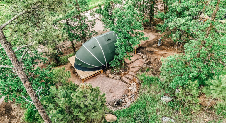 Aerial view of a modern glamping dome tent set on a raised wooden deck in a forested area, surrounded by trees, stone pathways, and natural greenery.