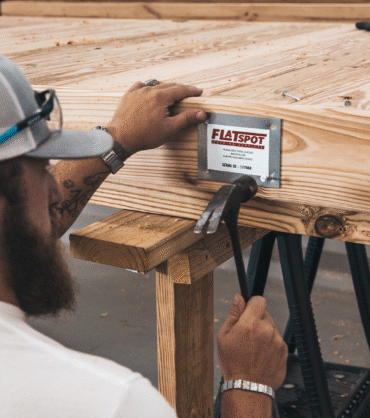 A craftsman works on assembling a modular wooden deck, securing a branded metal plate to the frame with nails and a hammer inside a woodworking workshop.