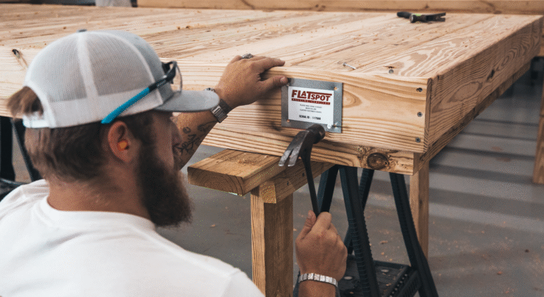A craftsman works on assembling a modular wooden deck, securing a branded metal plate to the frame with nails and a hammer inside a woodworking workshop.