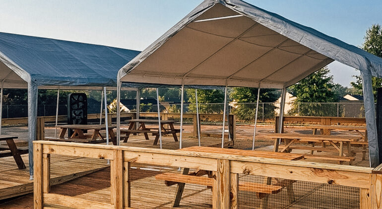 An outdoor seating area with wooden picnic tables placed under large canopy tents. The space is built on a wooden deck, enclosed by a light wooden fence, providing a shaded spot for gatherings or dining in a sunny open-air environment.
