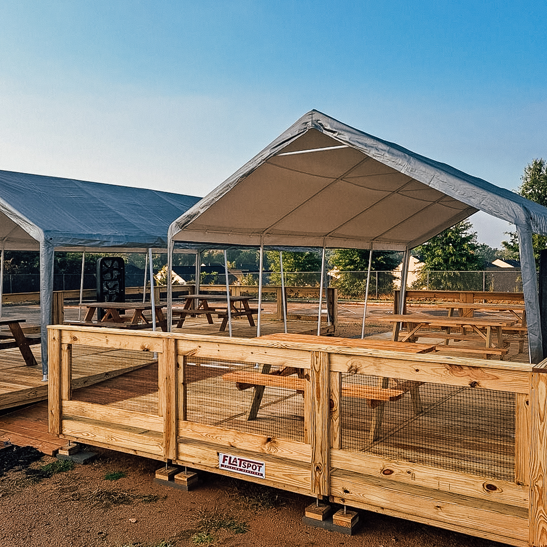 An outdoor seating area with wooden picnic tables placed under large canopy tents. The space is built on a wooden deck, enclosed by a light wooden fence, providing a shaded spot for gatherings or dining in a sunny open-air environment.