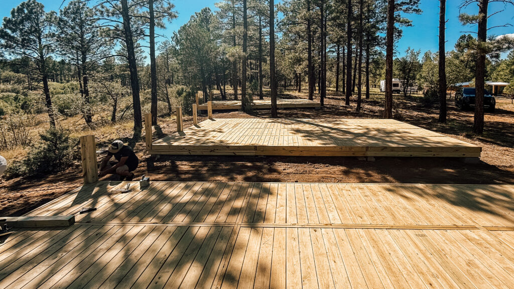 Workers assembling modular wooden decks in a forested campground area under clear blue skies, showing multiple elevated platforms in progress surrounded by tall pine trees.