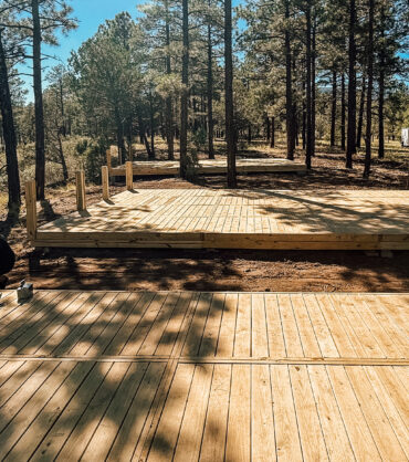 Workers assembling modular wooden decks in a forested campground area under clear blue skies, showing multiple elevated platforms in progress surrounded by tall pine trees.