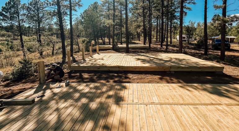 Workers assembling modular wooden decks in a forested campground area under clear blue skies, showing multiple elevated platforms in progress surrounded by tall pine trees.