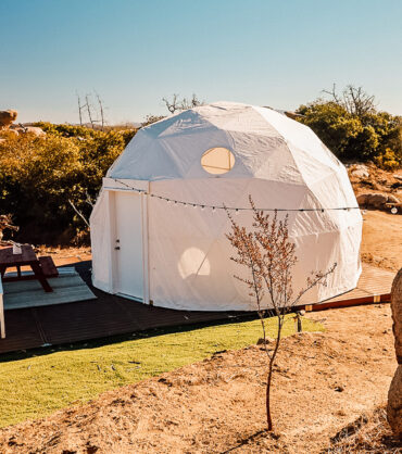 A modern white geodesic dome tent set on a wooden modular deck in a desert glamping site, surrounded by rocks, dry terrain, and scenic mountain views under a clear blue sky.