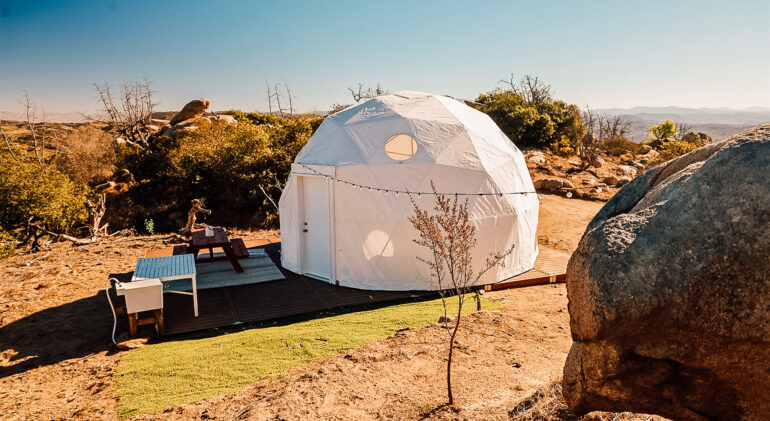 A modern white geodesic dome tent set on a wooden modular deck in a desert glamping site, surrounded by rocks, dry terrain, and scenic mountain views under a clear blue sky.