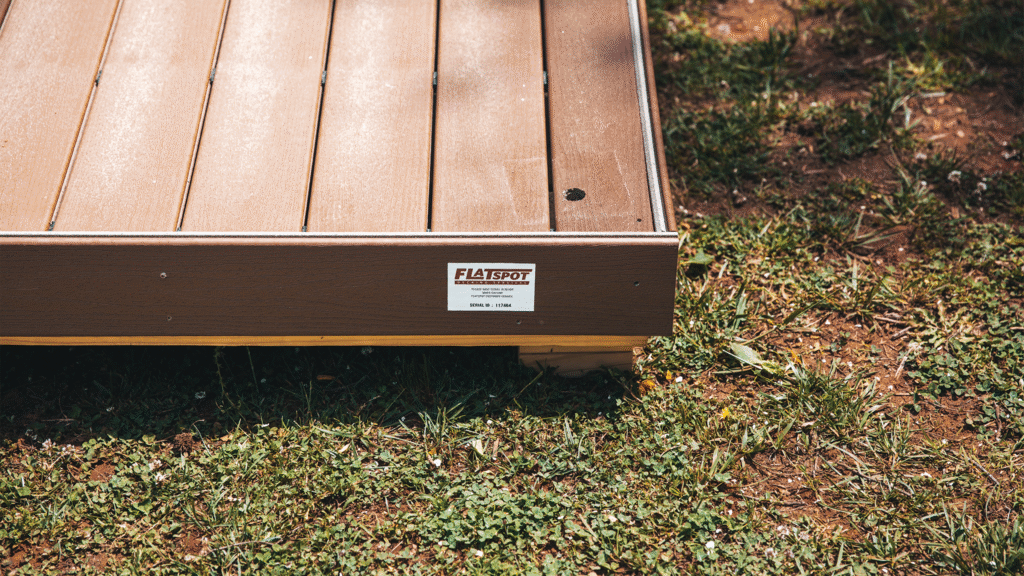 A close-up view of a raised wooden platform or deck placed on grass, showing brown composite decking boards with a small hole in one plank. A label on the side reads “FLATSPOT,” indicating the platform’s manufacturer. Sunlight highlights the texture of the boards and the surrounding patchy grass.