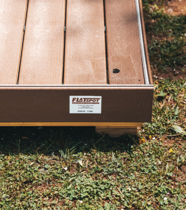 A close-up view of a raised wooden platform or deck placed on grass, showing brown composite decking boards with a small hole in one plank. A label on the side reads “FLATSPOT,” indicating the platform’s manufacturer. Sunlight highlights the texture of the boards and the surrounding patchy grass.