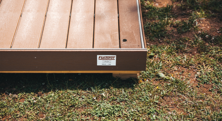 A close-up view of a raised wooden platform or deck placed on grass, showing brown composite decking boards with a small hole in one plank. A label on the side reads “FLATSPOT,” indicating the platform’s manufacturer. Sunlight highlights the texture of the boards and the surrounding patchy grass.