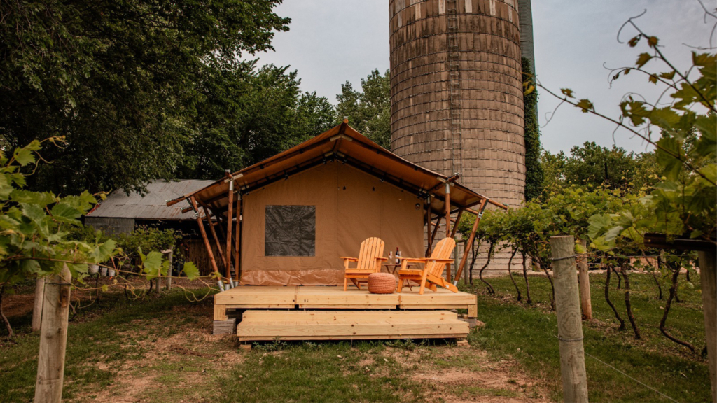A cozy glamping tent with a wooden deck and two Adirondack chairs sits among green vineyard rows, with a tall concrete silo and rustic farm buildings in the background.
