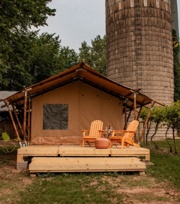 A cozy glamping tent with a wooden deck and two Adirondack chairs sits among green vineyard rows, with a tall concrete silo and rustic farm buildings in the background.