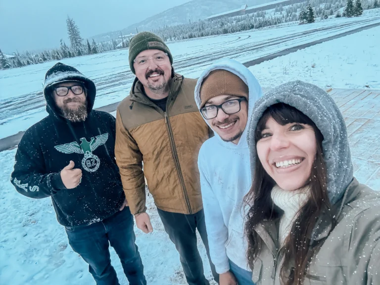 Four people bundled in winter clothing smile at the camera in a snowy outdoor setting, with mountains and trees in the background. The photo shows Joe Tim, the owner-operator of Cedar Sauna House together with the Flatspot Decking Services team.