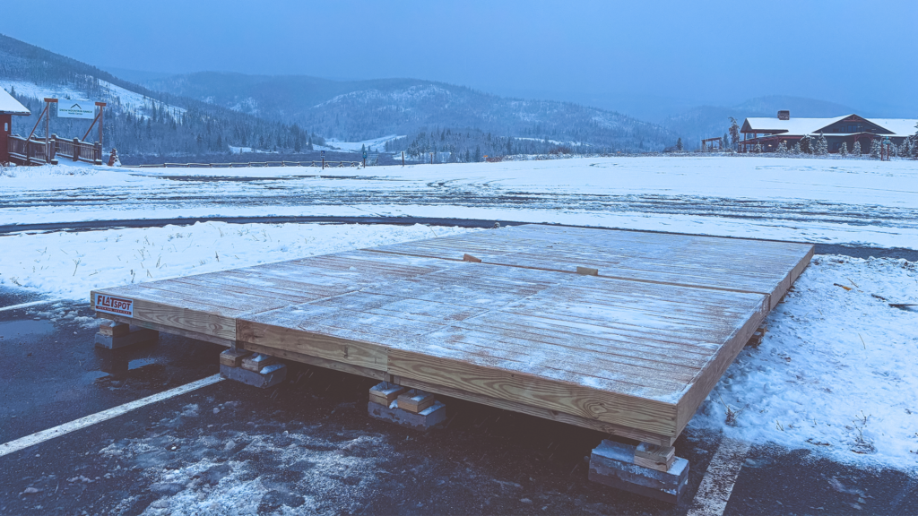 A wooden platform under construction sits on cinder blocks in a snowy parking lot, with snow-covered fields, trees, buildings, and mountains in the background.