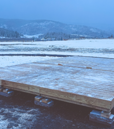 A wooden platform under construction sits on cinder blocks in a snowy parking lot, with snow-covered fields, trees, buildings, and mountains in the background.