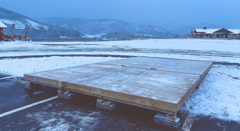 A wooden platform under construction sits on cinder blocks in a snowy parking lot, with snow-covered fields, trees, buildings, and mountains in the background.