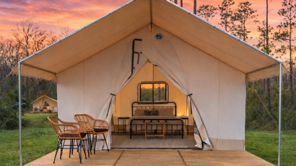A luxury glamping tent at sunset, featuring a wooden deck with two wicker chairs in the foreground. Inside the open canvas tent, there is a neatly made bed with a black metal frame and bedside tables. The background shows a wooded area under a vibrant orange and pink sky, with another similar tent visible in the distance.