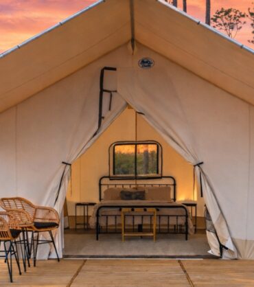 A luxury glamping tent at sunset, featuring a wooden deck with two wicker chairs in the foreground. Inside the open canvas tent, there is a neatly made bed with a black metal frame and bedside tables. The background shows a wooded area under a vibrant orange and pink sky, with another similar tent visible in the distance.