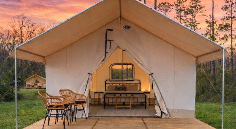 A luxury glamping tent at sunset, featuring a wooden deck with two wicker chairs in the foreground. Inside the open canvas tent, there is a neatly made bed with a black metal frame and bedside tables. The background shows a wooded area under a vibrant orange and pink sky, with another similar tent visible in the distance.