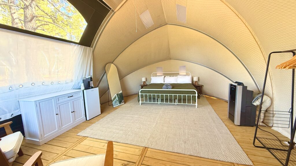 The interior of a luxury glamping dome featuring a wooden floor, a large bed with a green comforter, and a neutral-toned rug. The space is furnished with a white dresser, a mini-fridge, a full-length mirror, and a portable air conditioner. Natural light enters through a large window and a transparent ceiling section revealing trees outside.