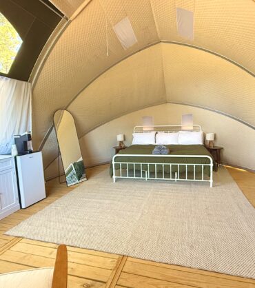 The interior of a luxury glamping dome featuring a wooden floor, a large bed with a green comforter, and a neutral-toned rug. The space is furnished with a white dresser, a mini-fridge, a full-length mirror, and a portable air conditioner. Natural light enters through a large window and a transparent ceiling section revealing trees outside.