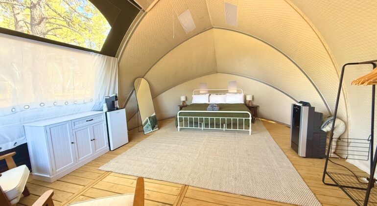 The interior of a luxury glamping dome featuring a wooden floor, a large bed with a green comforter, and a neutral-toned rug. The space is furnished with a white dresser, a mini-fridge, a full-length mirror, and a portable air conditioner. Natural light enters through a large window and a transparent ceiling section revealing trees outside.