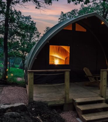 A cozy glamping pod with a wooden deck and two lounge chairs at dusk, featuring a warm glowing interior and an outdoor stone fire pit nestled in a lush green forest setting.