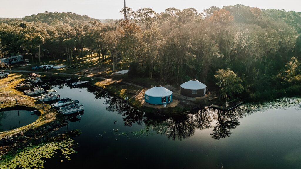 Aerial view of a lakeside glamping resort featuring two modern yurts, a boat dock with several pontoon boats, and a lush forest at sunset.