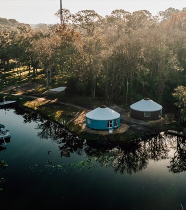 Aerial view of a lakeside glamping resort featuring two modern yurts, a boat dock with several pontoon boats, and a lush forest at sunset.