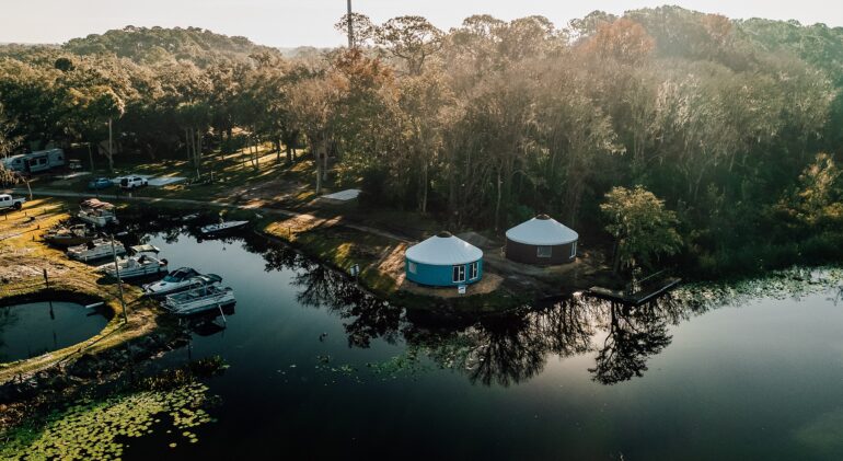 Aerial view of a lakeside glamping resort featuring two modern yurts, a boat dock with several pontoon boats, and a lush forest at sunset.