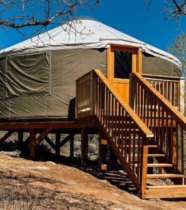 A detailed photograph of a modern yurt with a dark olive-green canvas body and a light-colored domed roof, set on an elevated wooden platform on a dry, brushy hillside. A full wooden deck and a set of wooden stairs with a banister lead up to a glass-paneled wooden front door. Bare tree branches frame the top of the yurt against a clear blue sky. The background shows arid hills and scrubland.
