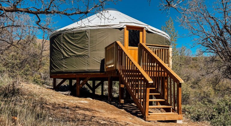 A detailed photograph of a modern yurt with a dark olive-green canvas body and a light-colored domed roof, set on an elevated wooden platform on a dry, brushy hillside. A full wooden deck and a set of wooden stairs with a banister lead up to a glass-paneled wooden front door. Bare tree branches frame the top of the yurt against a clear blue sky. The background shows arid hills and scrubland.