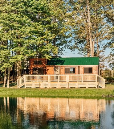 A wooden cabin with a green roof and a large deck, nestled in the trees and reflecting in a calm pond.