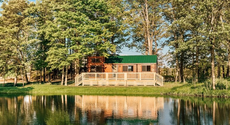 A wooden cabin with a green roof and a large deck, nestled in the trees and reflecting in a calm pond.