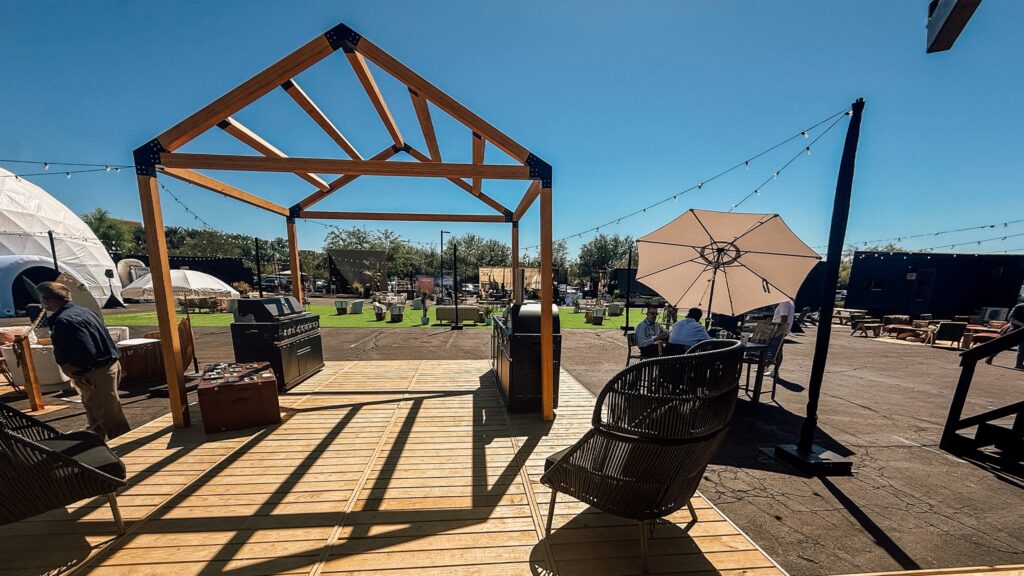 An outdoor event and lounge space on a sunny day. In the foreground, a wooden deck features an open, house-shaped timber frame structure, a barbecue grill, and a black wicker chair. In the background, there is an asphalt lot with patches of artificial grass, a white geodesic dome tent, string lights, dark modular container structures, and people relaxing at a table under a white umbrella.
