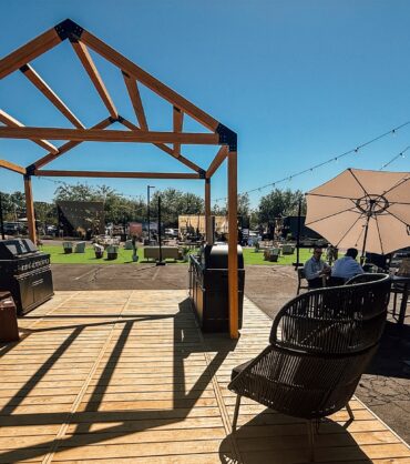 An outdoor event and lounge space on a sunny day. In the foreground, a wooden deck features an open, house-shaped timber frame structure, a barbecue grill, and a black wicker chair. In the background, there is an asphalt lot with patches of artificial grass, a white geodesic dome tent, string lights, dark modular container structures, and people relaxing at a table under a white umbrella.