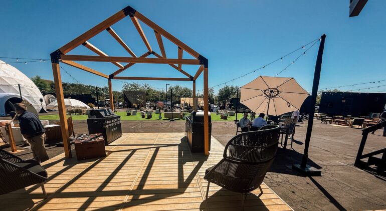 An outdoor event and lounge space on a sunny day. In the foreground, a wooden deck features an open, house-shaped timber frame structure, a barbecue grill, and a black wicker chair. In the background, there is an asphalt lot with patches of artificial grass, a white geodesic dome tent, string lights, dark modular container structures, and people relaxing at a table under a white umbrella.