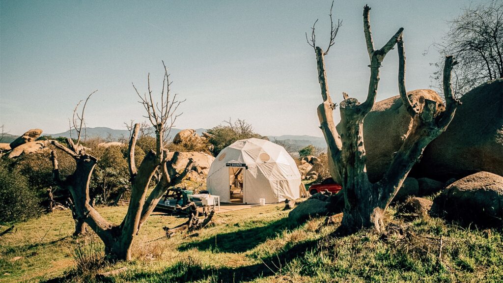 A white geodesic glamping dome with an open entrance, situated in a rugged, sunlit landscape. The dome, bearing a small sign above the door that reads 'THE CROWN', sits on a wooden deck. The scene is framed by two large, bare, dead tree trunks in the foreground. The surrounding area features sparse green grass, large boulders, and a red vehicle partially visible behind the dome, with rolling mountains in the distance under a clear blue sky.